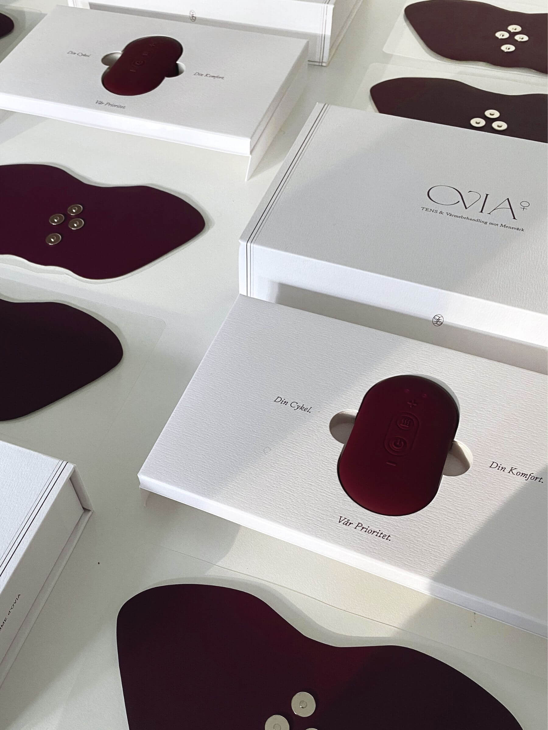 Flera vita förpackningar som innehåller Ovia⁺, en burgundyfärgad oval TENS-inspirerad wellness-teknologi och enhet, placerad i en elegant och minimalistisk förpackning.