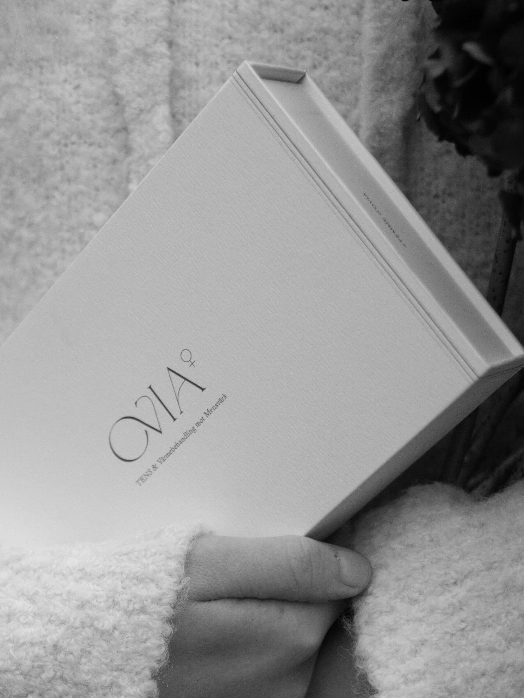 En svartvit närbild av en kvinna med ljus stickad tröja håller en Ovia⁺ i sin famn, placerad i en elegant och minimalistisk förpackning.
