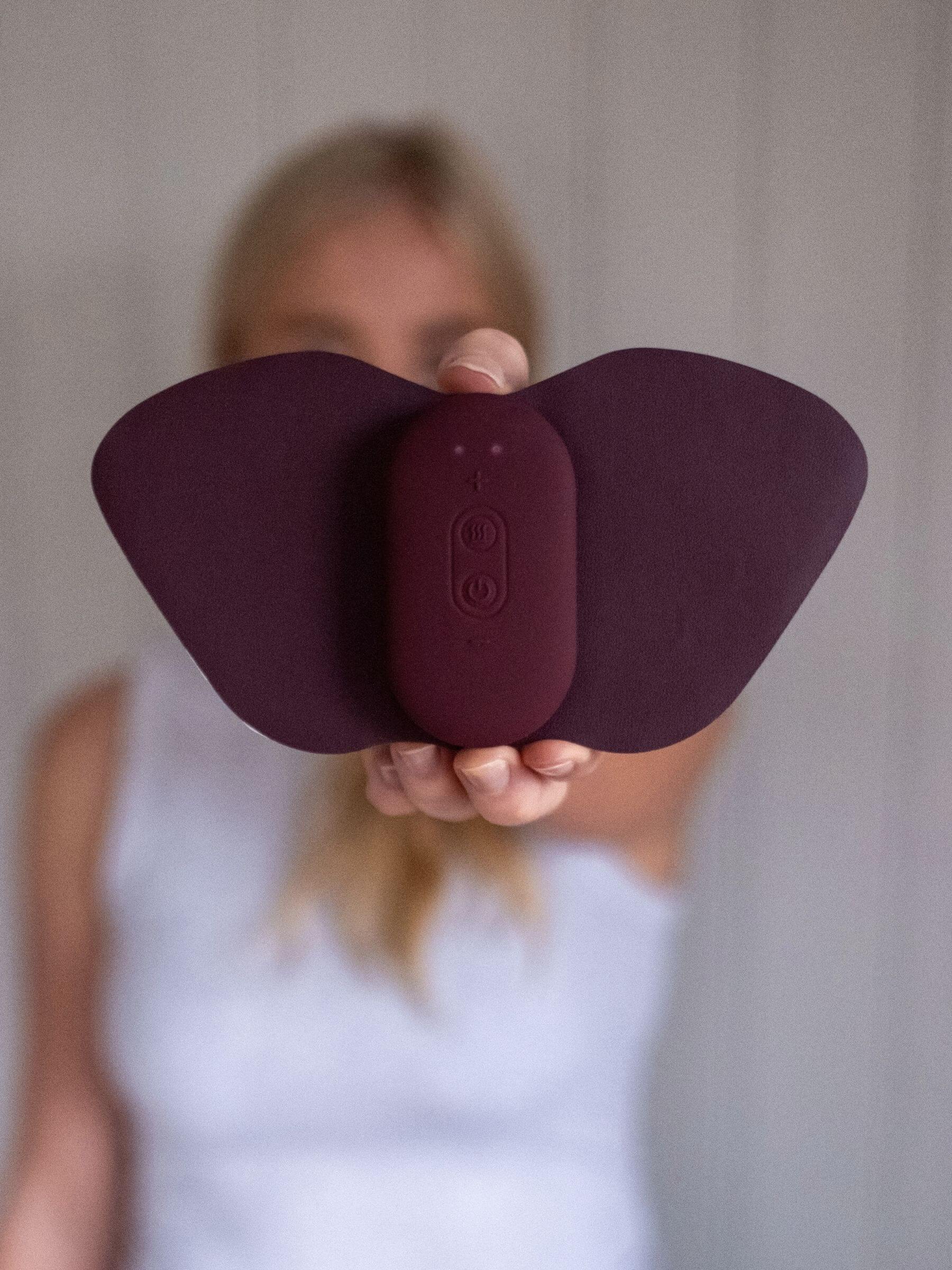 En kvinna håller en Ovia⁺ i handen, representerar TENS-baserad egenvård med värme och sträcker ut den mot kameran. Bakgrunden är mjukt suddig.