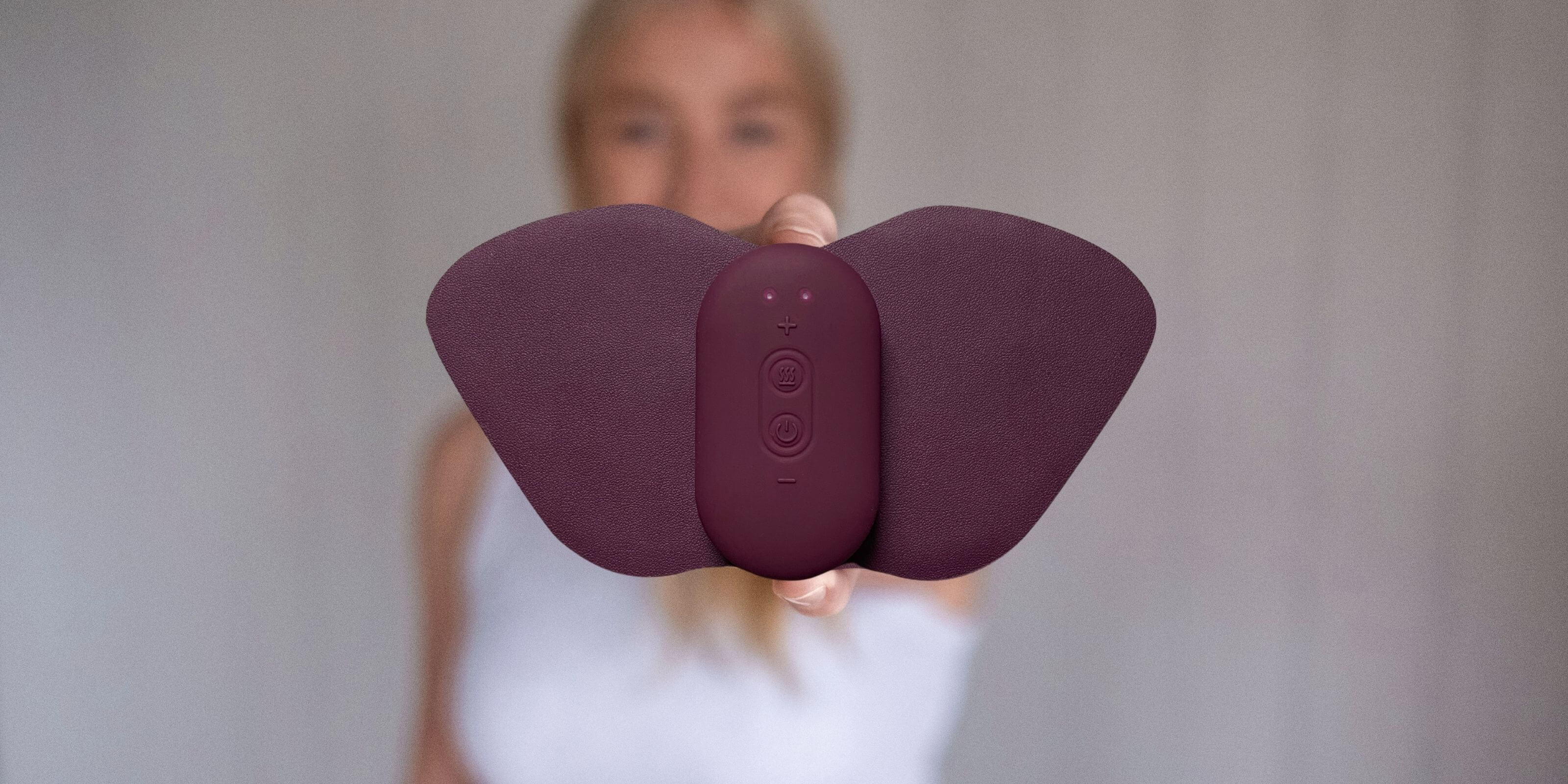 En kvinna håller en Ovia⁺ i handen, representerar TENS-baserad egenvård med värme och sträcker ut den mot kameran. Bakgrunden är mjukt suddig.