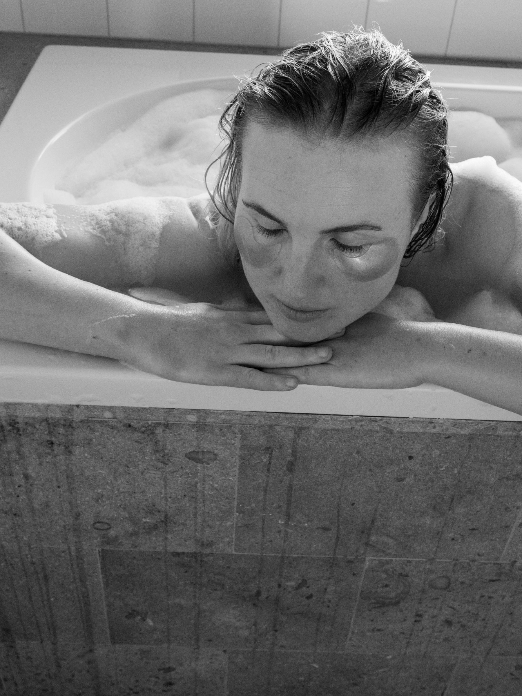 En svartvit närbild av en kvinna som sitter i ett badkar och lutar sig över kanten. Hon har en ansiktsmask, stämningen är avslappnad och förmedlar en känsla av egenvård.