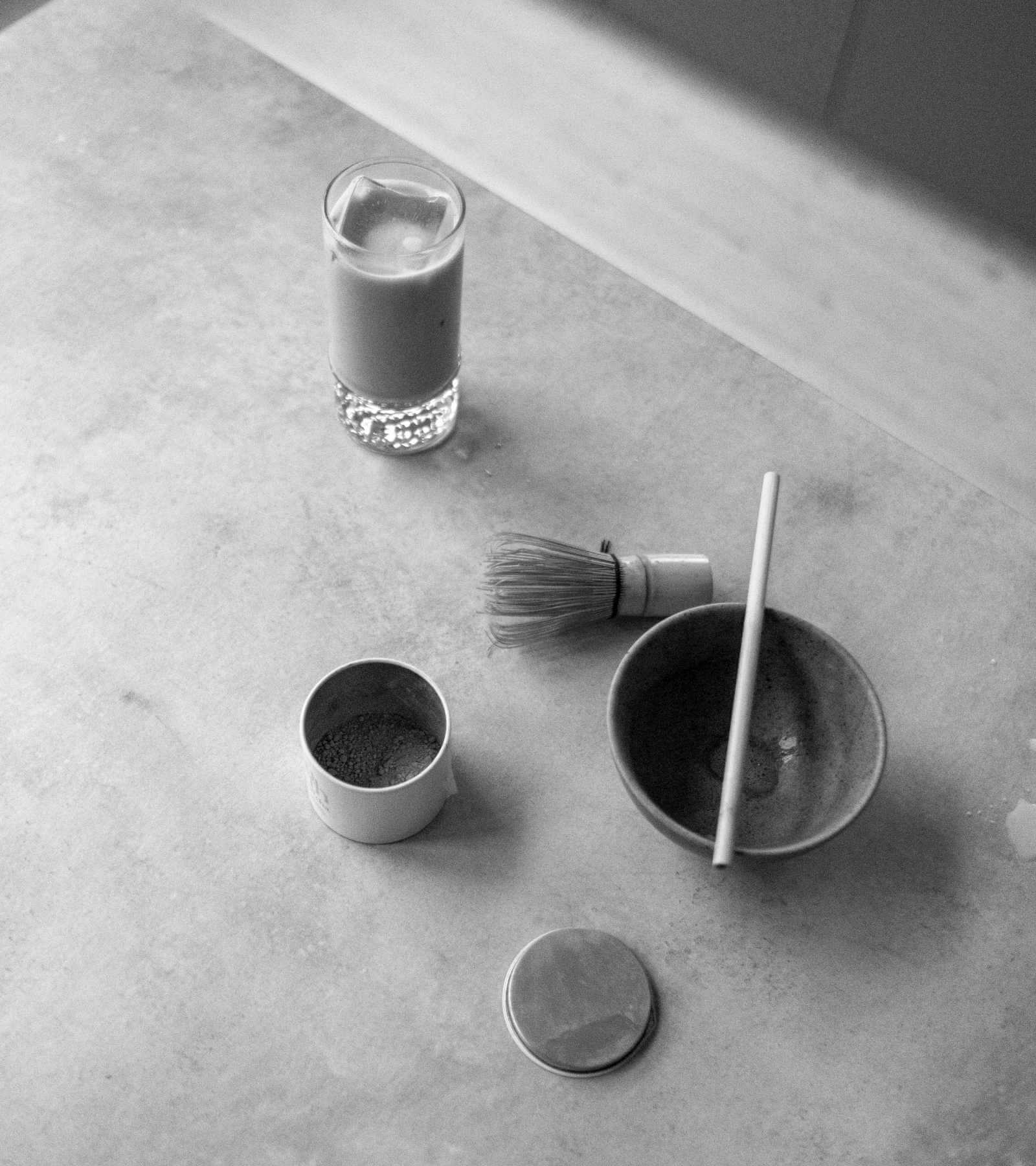 Svartvit bild på en minimalistisk matcha uppsättning: ett glas med matcha, en visp, en matchaskål med omrörare och en öppen behållare med matchapulver på en texturerad yta. Lugnt och fridfullt.