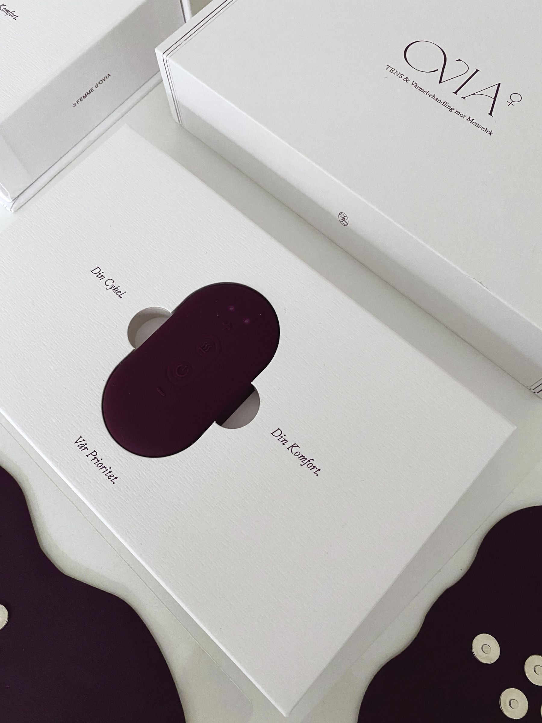 En vit förpackning som innehåller Ovia⁺, en burgundyfärgad oval TENS-inspirerad wellness-teknologi och enhet, placerad i en elegant och minimalistisk förpackning.
