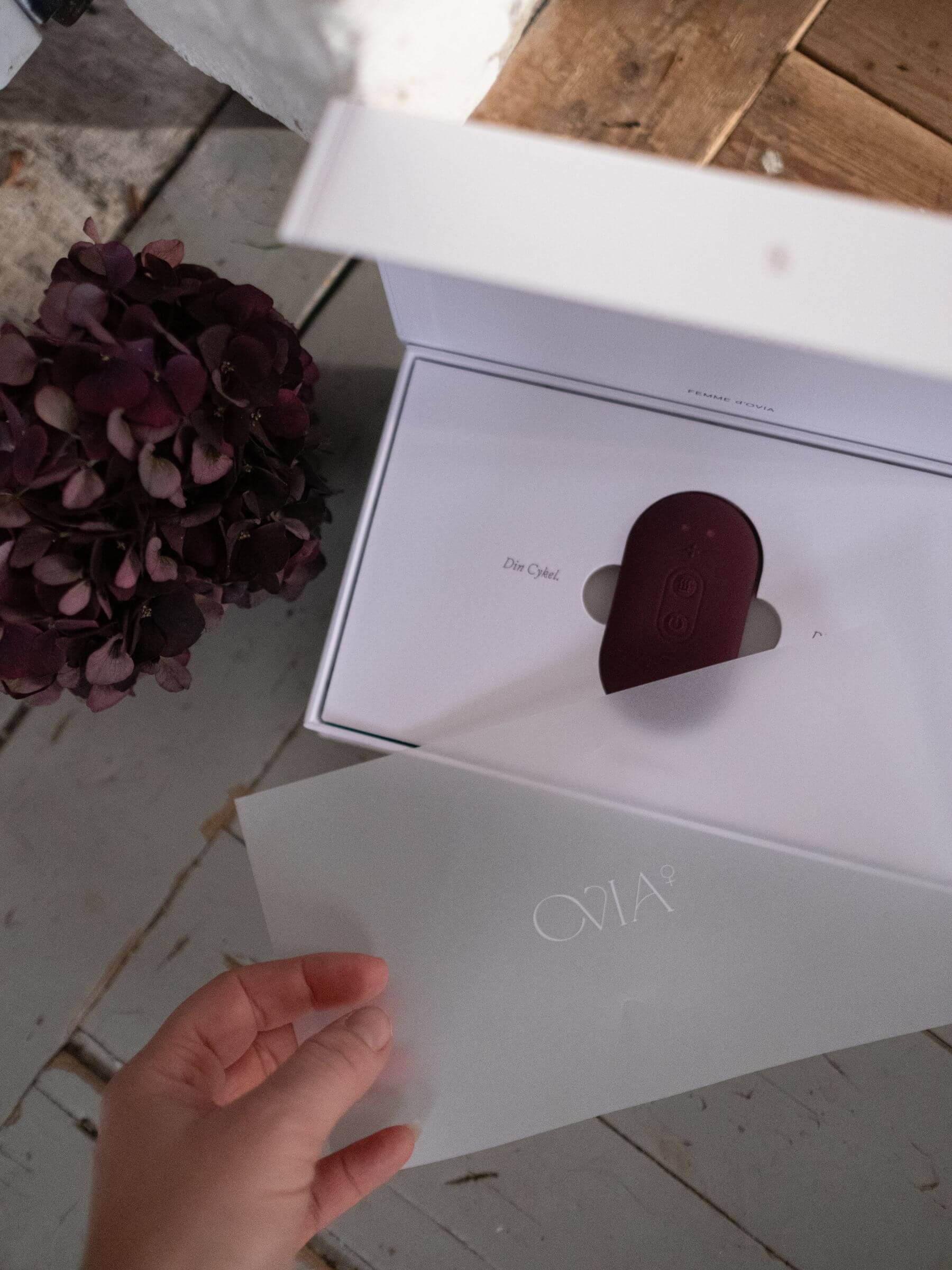 En hand lyfter upp ett frostat papper och avsljar en vit förpackning som innehåller Ovia⁺, en burgundyfärgad oval TENS-inspirerad wellness-teknologi och enhet, placerad i en elegant och minimalistisk förpackning.