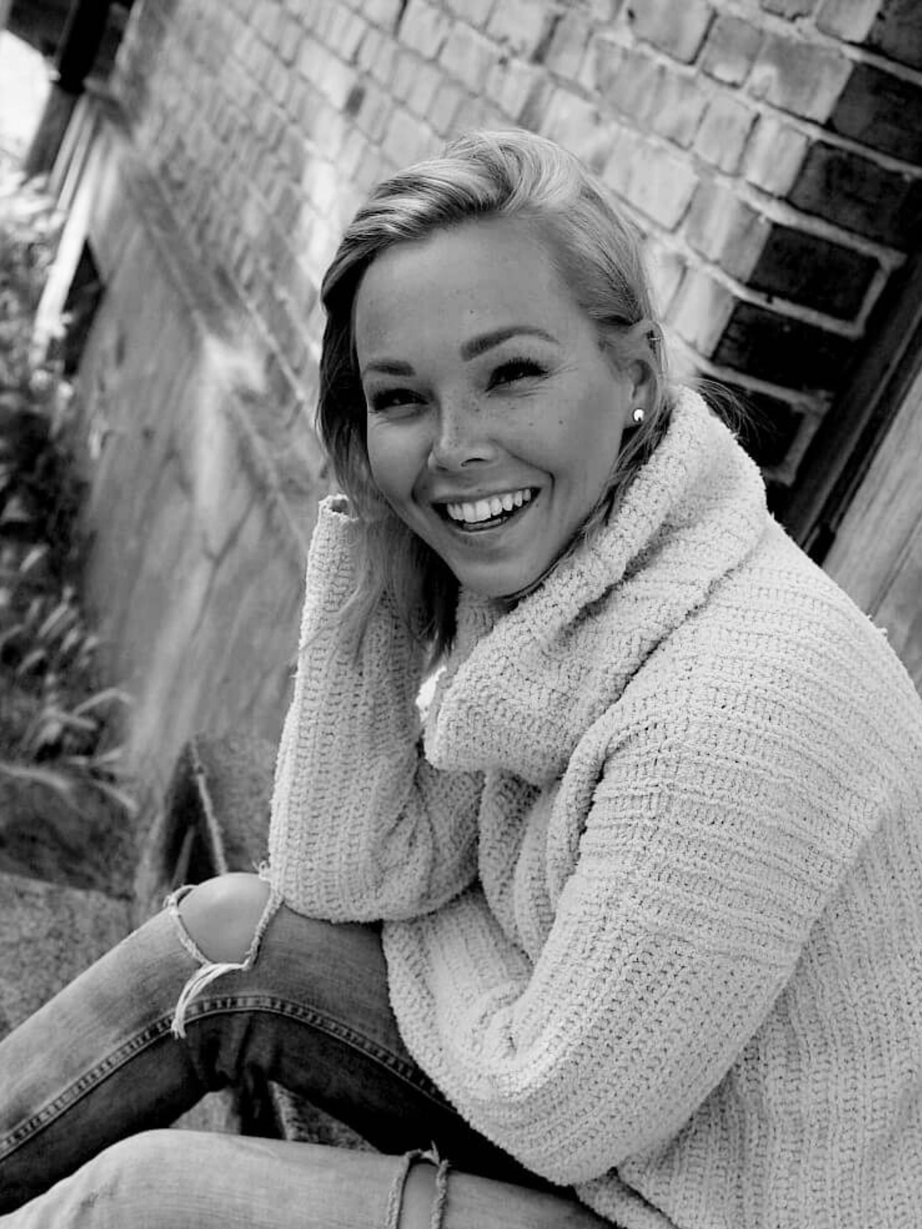 En svartvit bild av en kvinna i slitna jeans och stickad tröja som sitter på en trappa utanför ett tegelhus