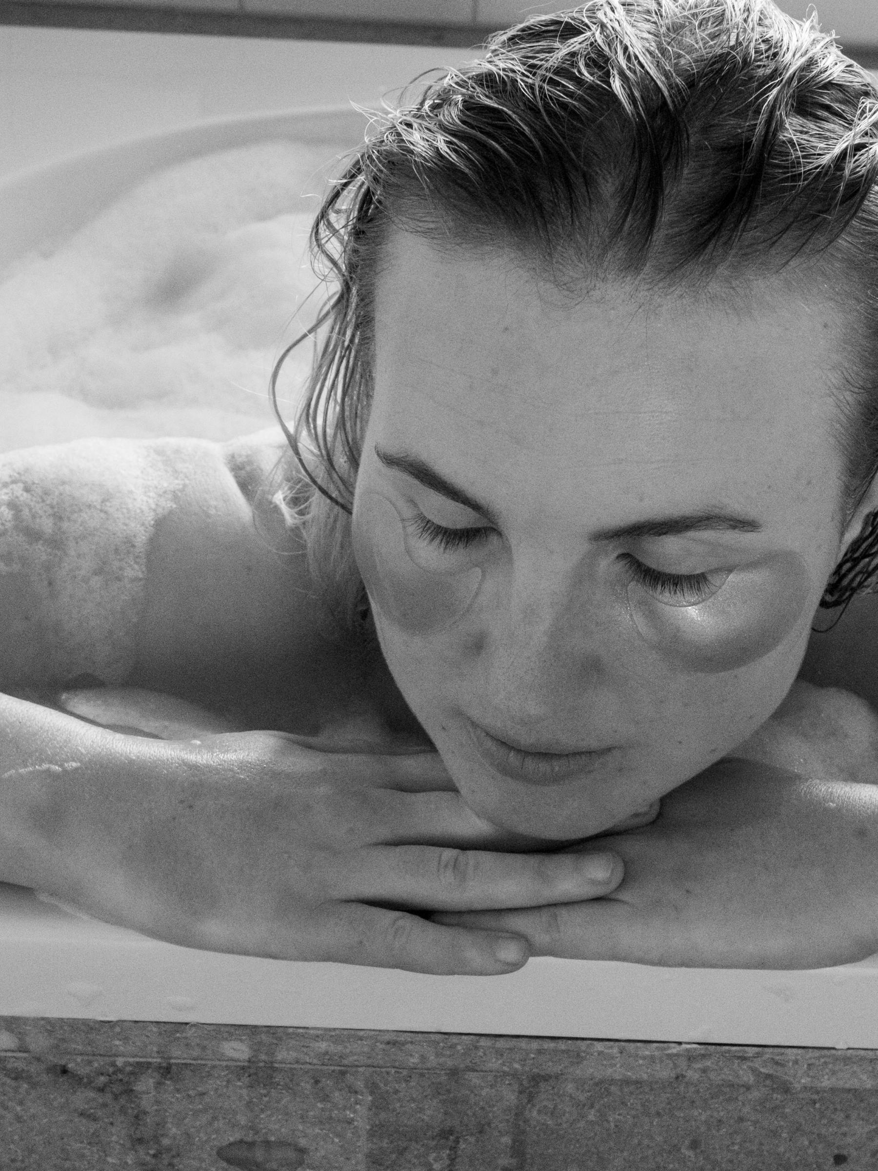 En svartvit närbild av en kvinna som sitter i ett badkar och lutar sig över kanten. Hon har en ansiktsmask, stämningen är avslappnad och förmedlar en känsla av egenvård. 