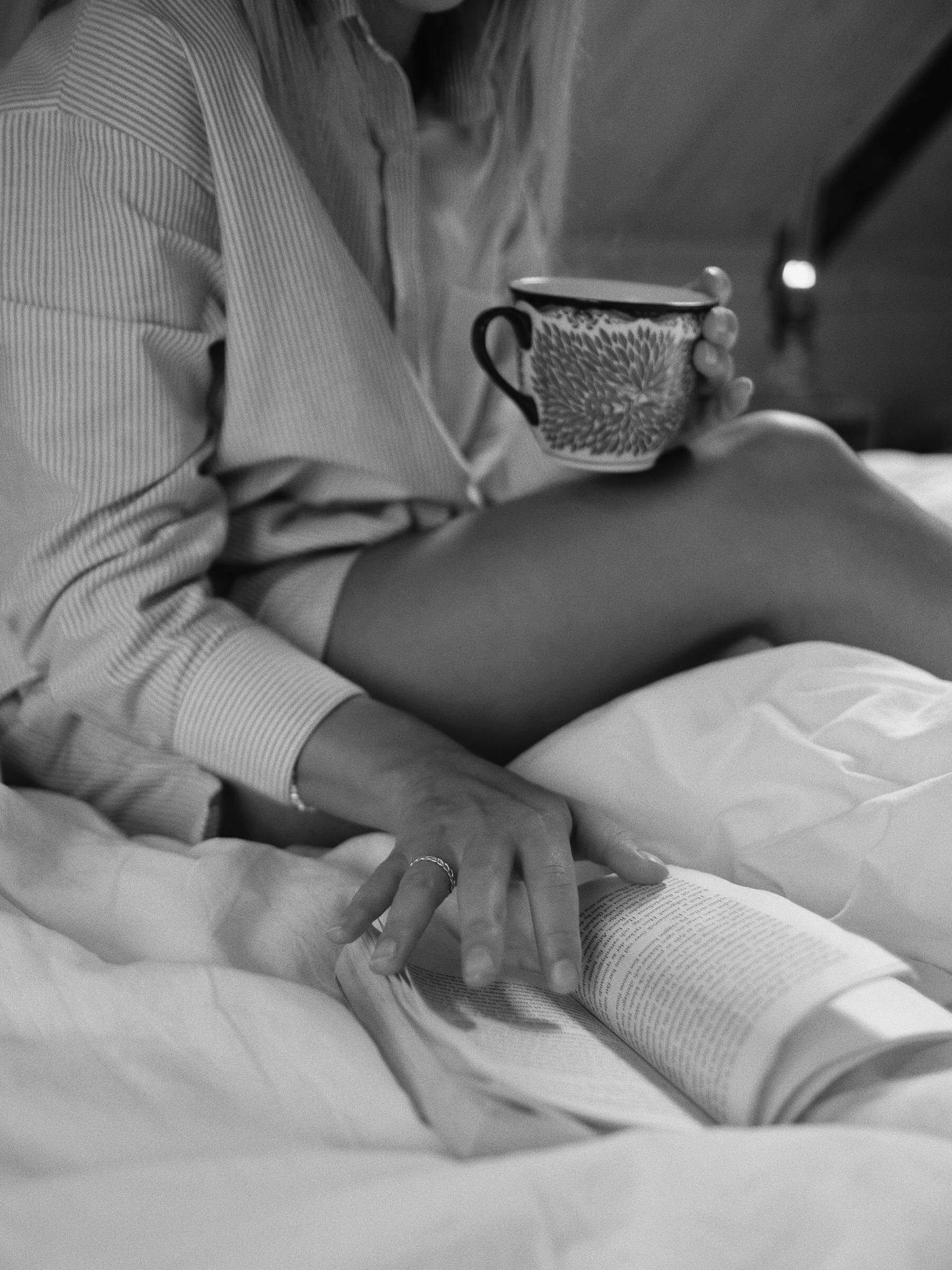 En kvinna i randig skjorta sitter på en säng med en mönstrad mugg i handen och rör försiktigt vid en uppslagen bok. Scenen förmedlar en mysig, avslappnad morgonkänsla.