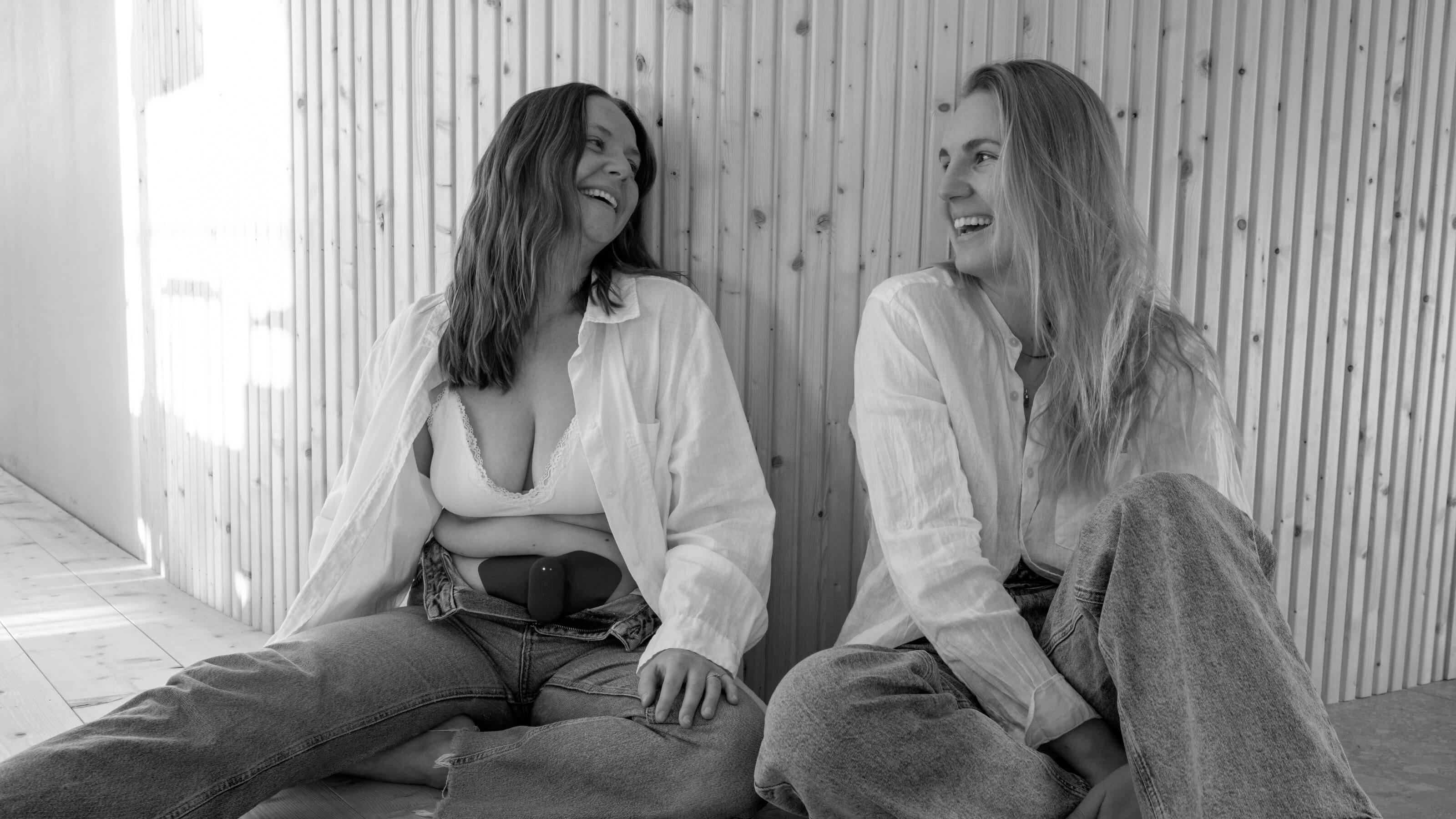 En svartvit bild av två kvinnor sitter på golvet och bär vita skjortor och jeans.  De använder Ovia + mot menssmärta och är glada för att de upplever lindring. Stämningen är glad och avslappnad. 