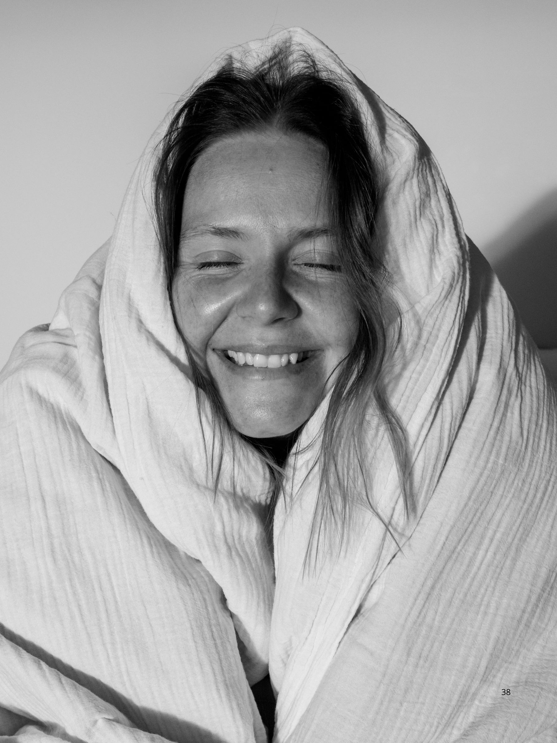 En svartvit bild av en glad kvinna invirad i ett ljust täcke. Hon ler och stämningen är genuin, avslappnad och positiv.  