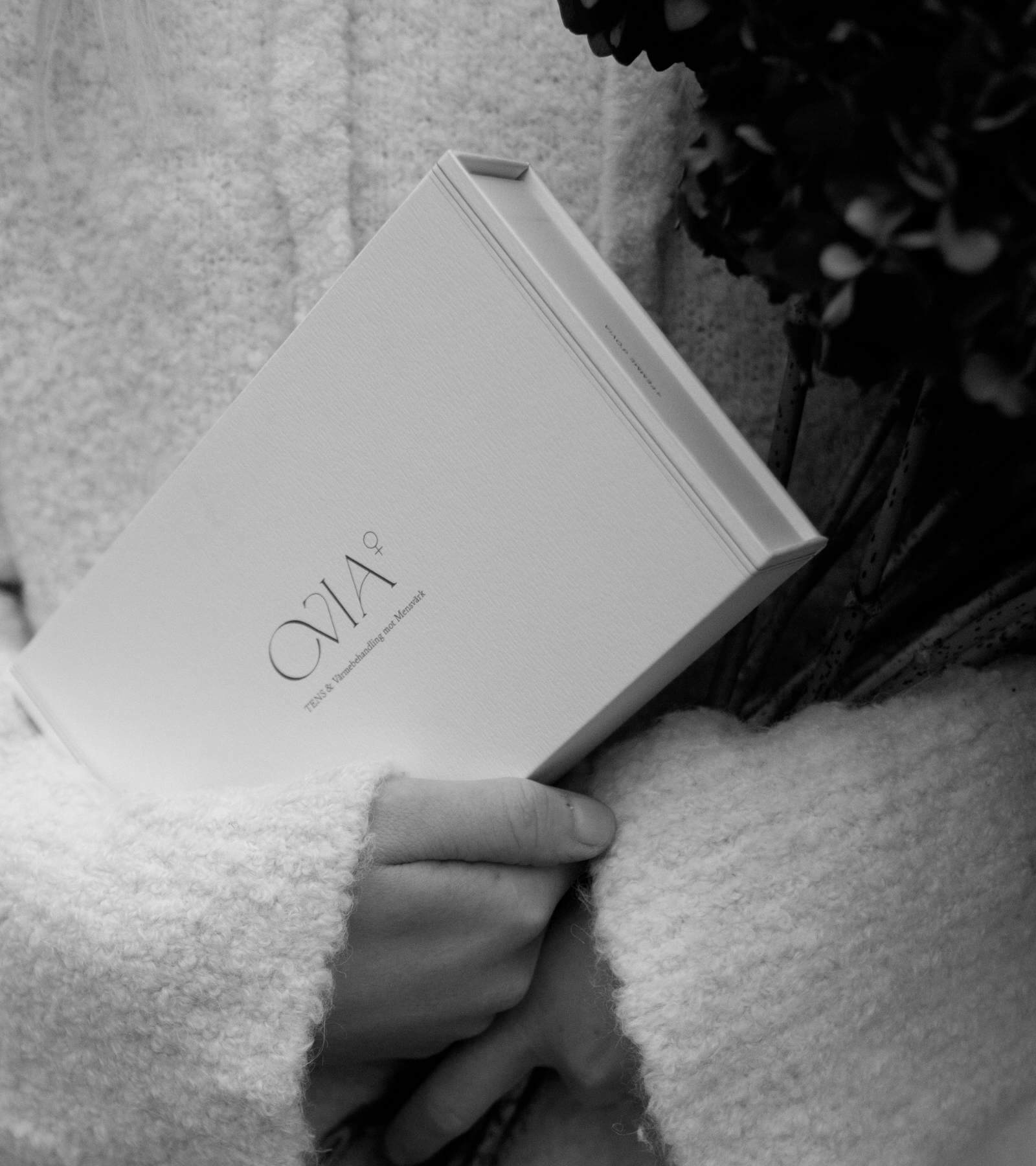 En svartvit närbild av en kvinna med ljus stickad tröja håller en Ovia⁺ i sin famn,  placerad i en elegant och minimalistisk förpackning.