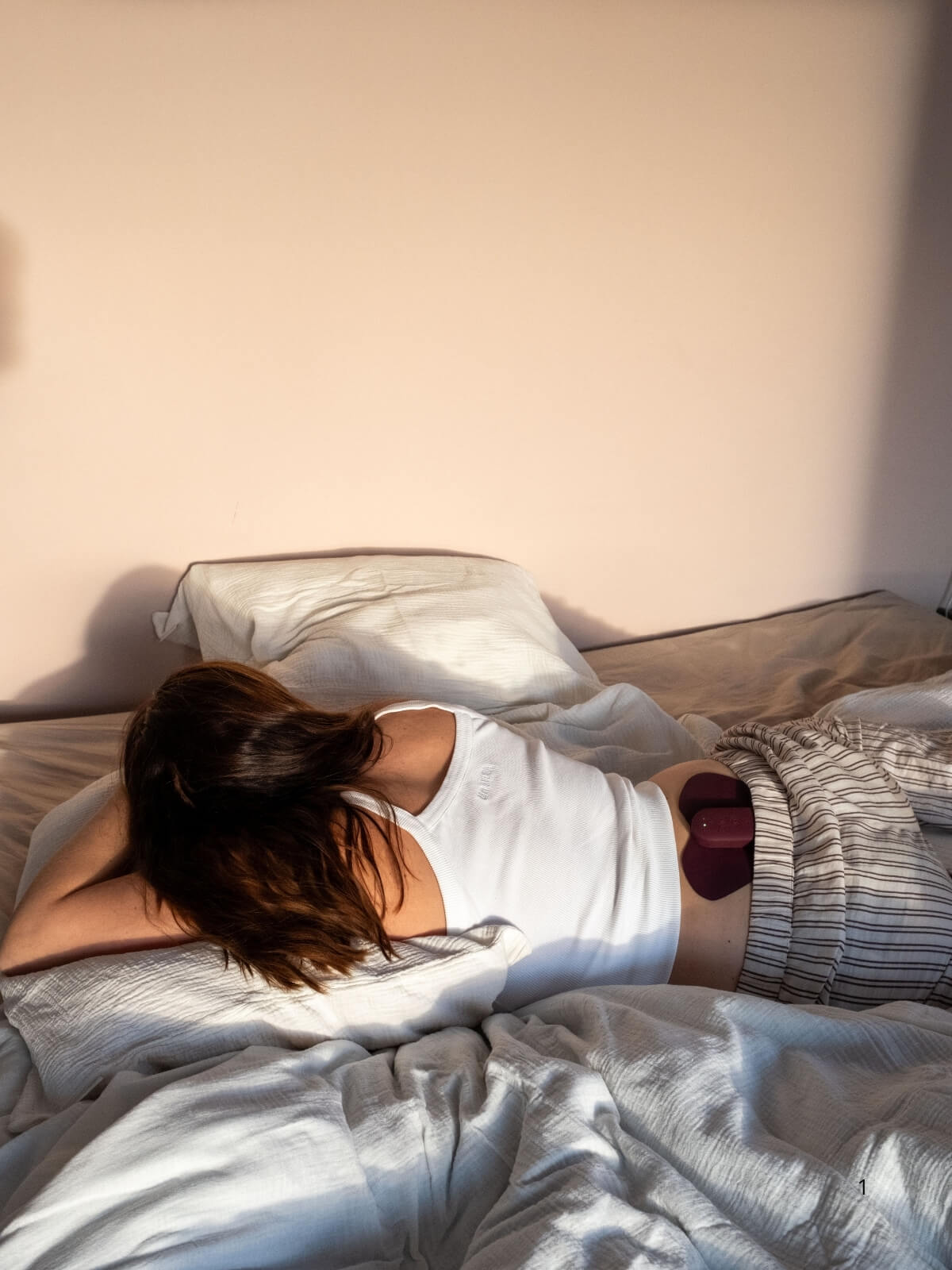 En kvinna som bär vitt linne och randiga pyamasbyxor ligger avslappnat i en säng på magen. Hon bär Ovia, en TENS-inspirerat apparat mot smärta på ländryggen. Stämningen är avslappnad och mysig. 