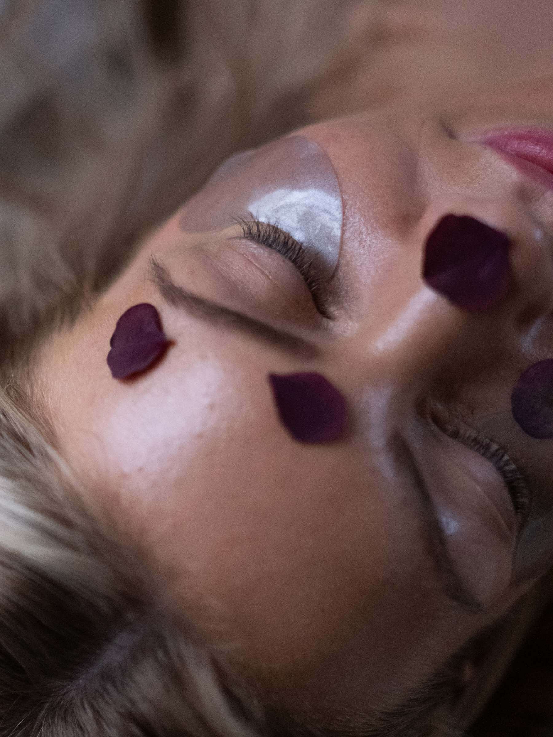 En närbild av en kvinnans ansikte sett snett ovanifrån med blomblad på ansiktet och ansiktsmask. Bilden utstrålar lugn och  harmoni. 